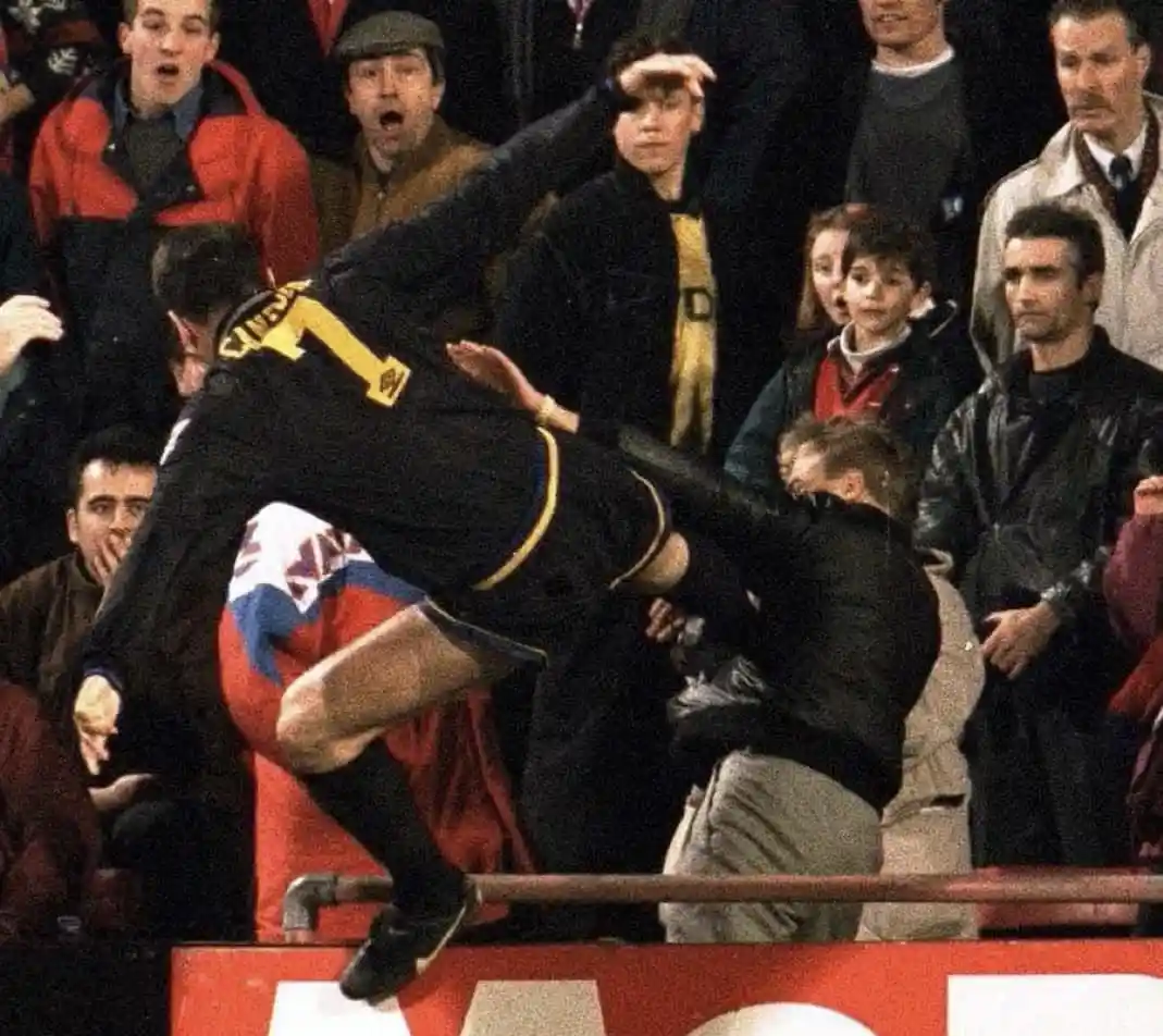 A soccer player in a black jersey with the number 1 is mid-air, leaping over a barrier into the stands, while a spectator in…