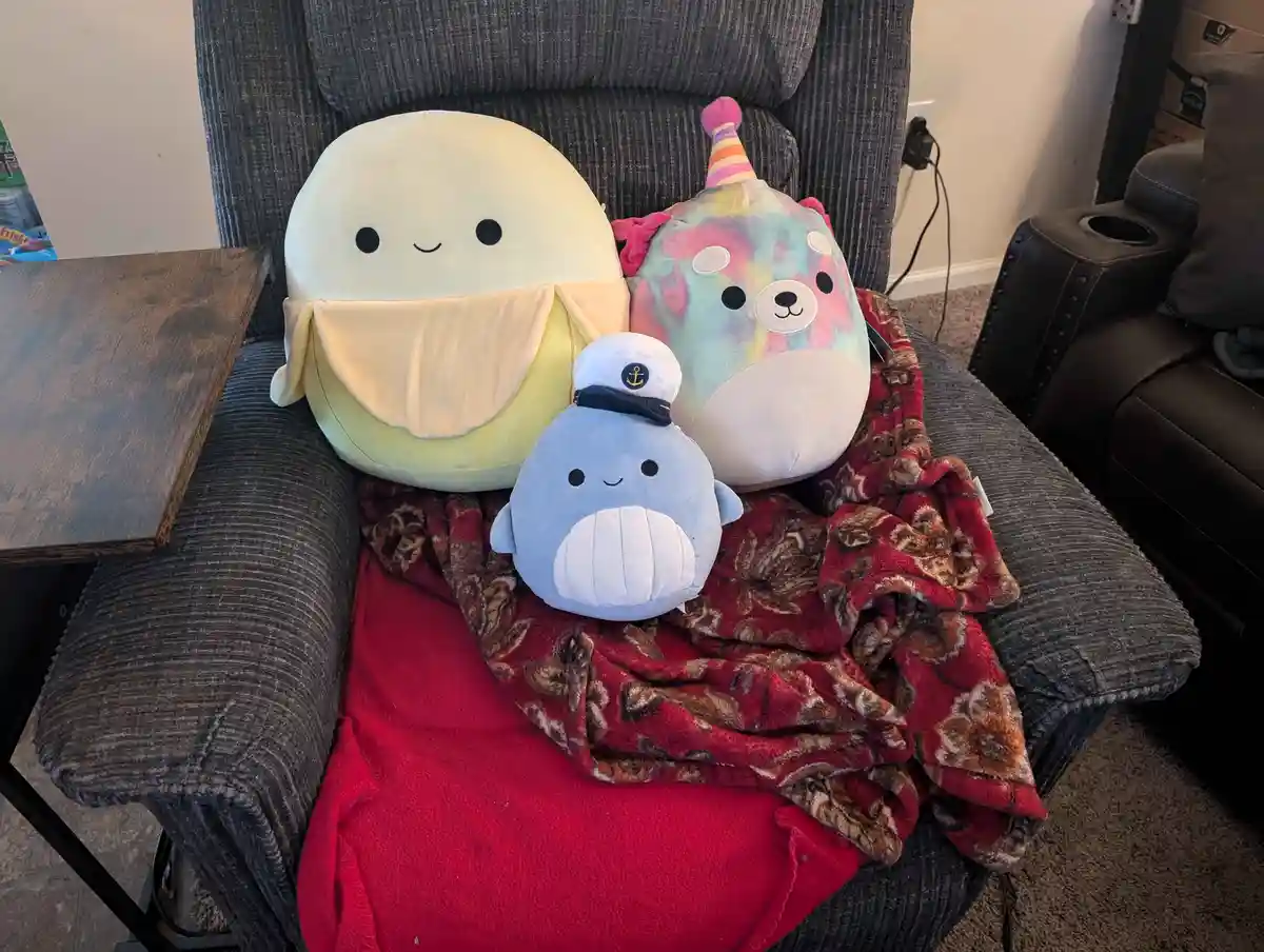 Three Squishmallows plush toys are sitting together on a gray recliner chair covered with blankets. The largest is a pale…