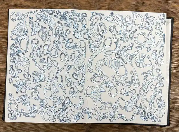 An open sketchbook lies on a wooden surface, filled with intricate, abstract blue line drawings that resemble tangled, wavy…