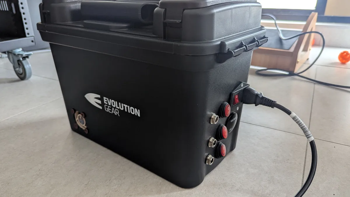 A black, rugged-looking portable power station branded 'EVOLUTION GEAR' sits on a tiled floor, featuring multiple red power…