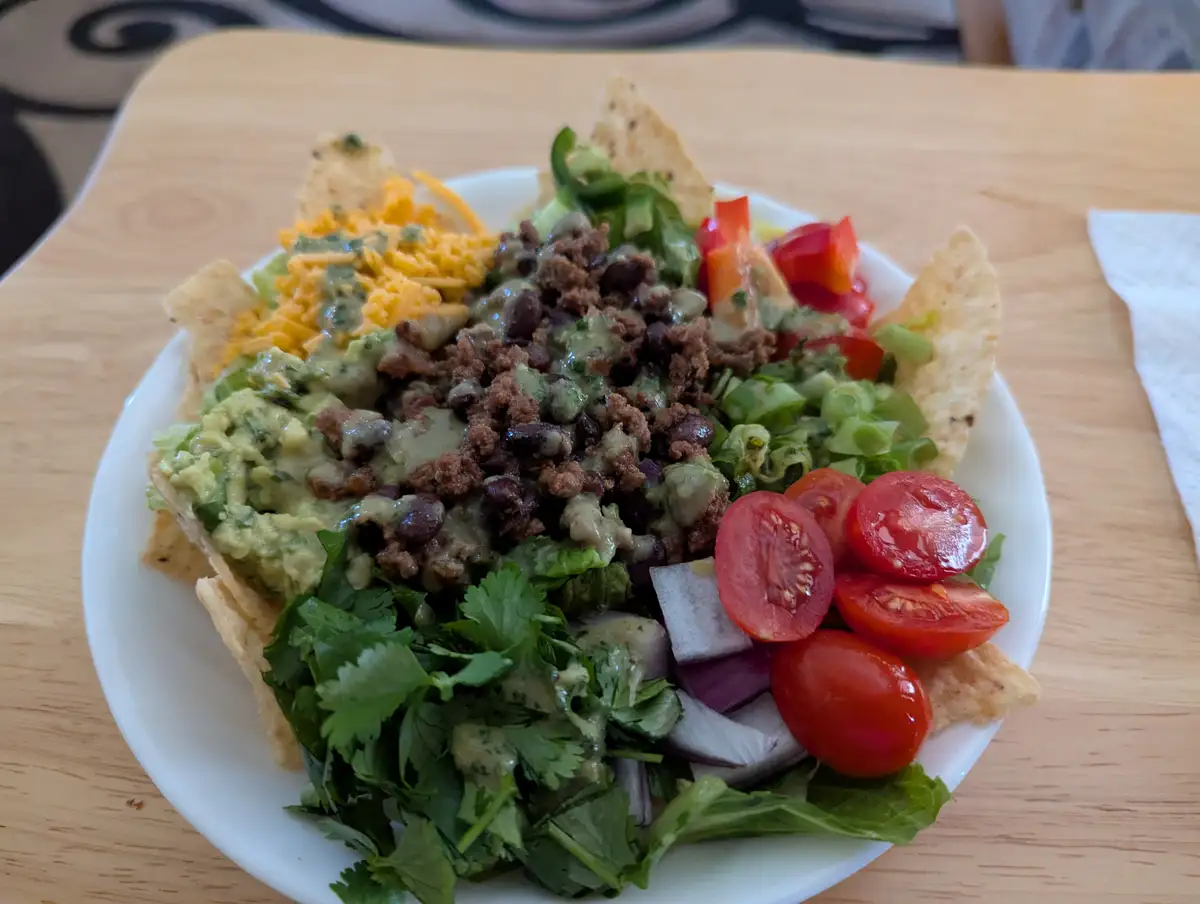 A white plate holds a colorful nacho bowl filled with tortilla chips, shredded cheese, guacamole, seasoned ground meat…