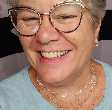 A close-up photo of a smiling older woman with short gray hair, wearing large tortoiseshell-framed glasses and a silver…