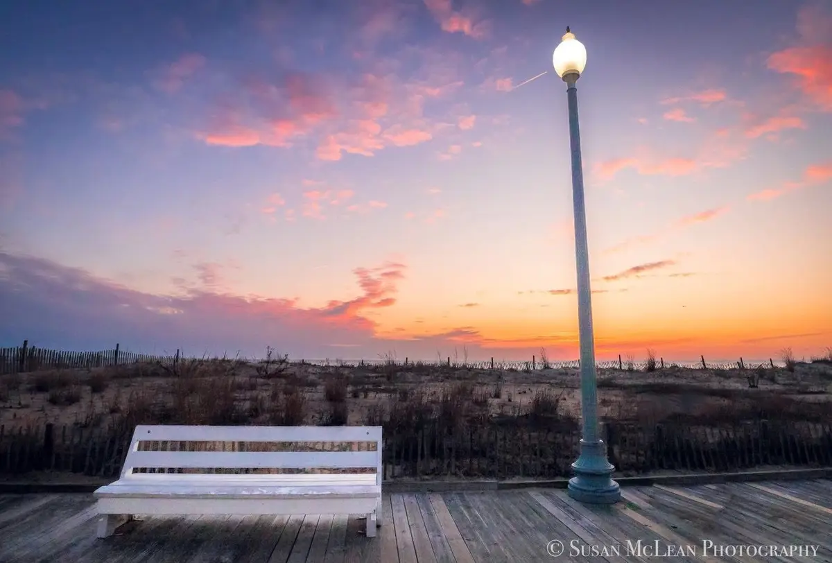 A serene beach scene at sunset with a white wooden bench on a boardwalk, next to a lit lamppost, under a sky painted with…