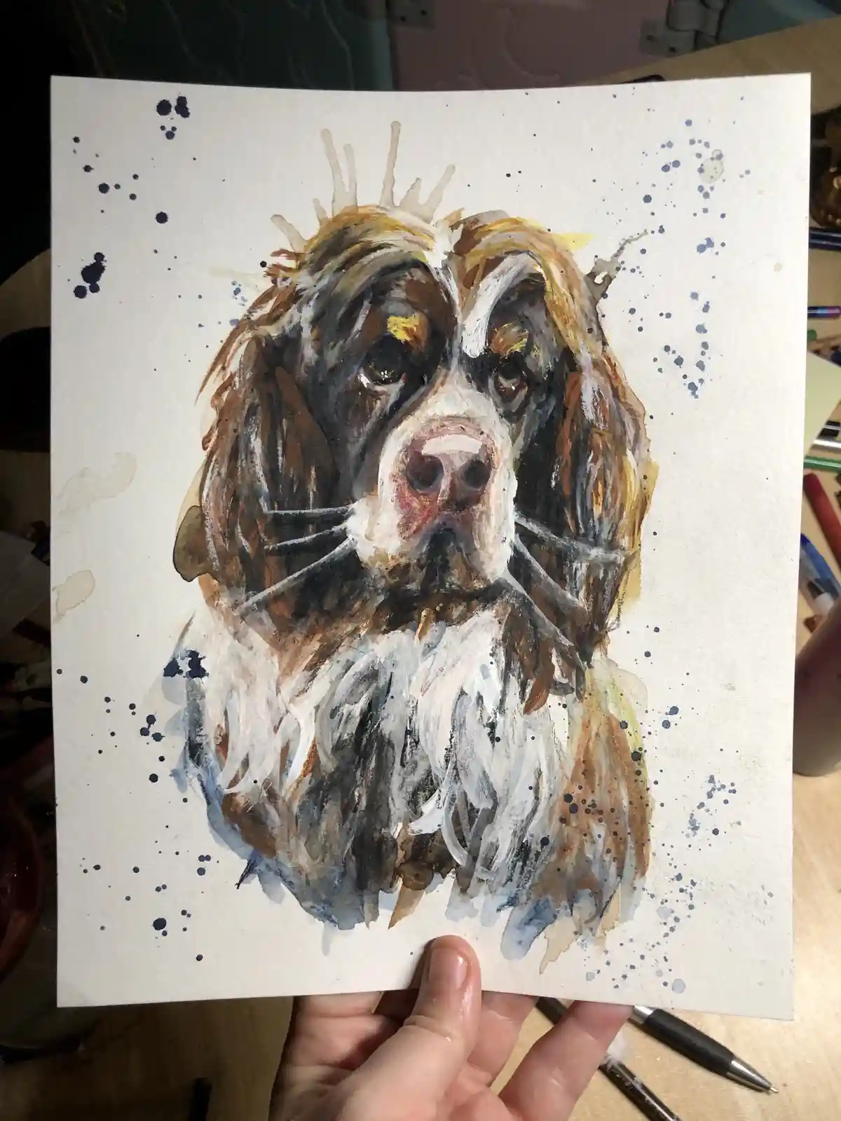 A hand holds up a watercolor painting of a dog with brown, white, and black fur, featuring a crown-like splash of paint on…