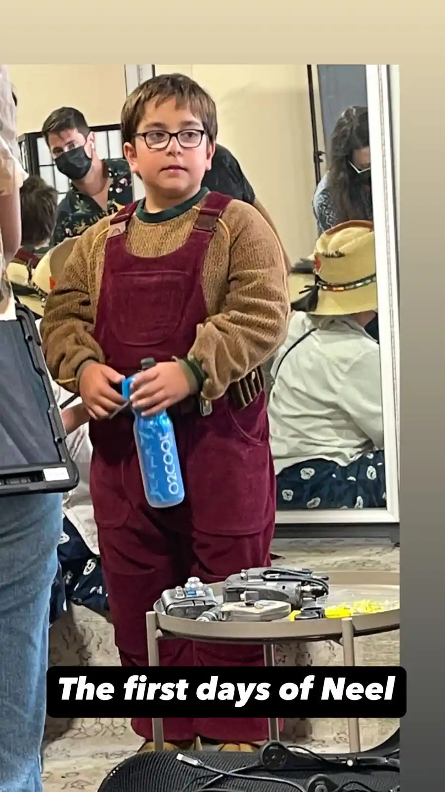 A young boy with glasses, wearing maroon overalls over a brown sweater, stands holding a blue 'O2COOL' water bottle. He has…