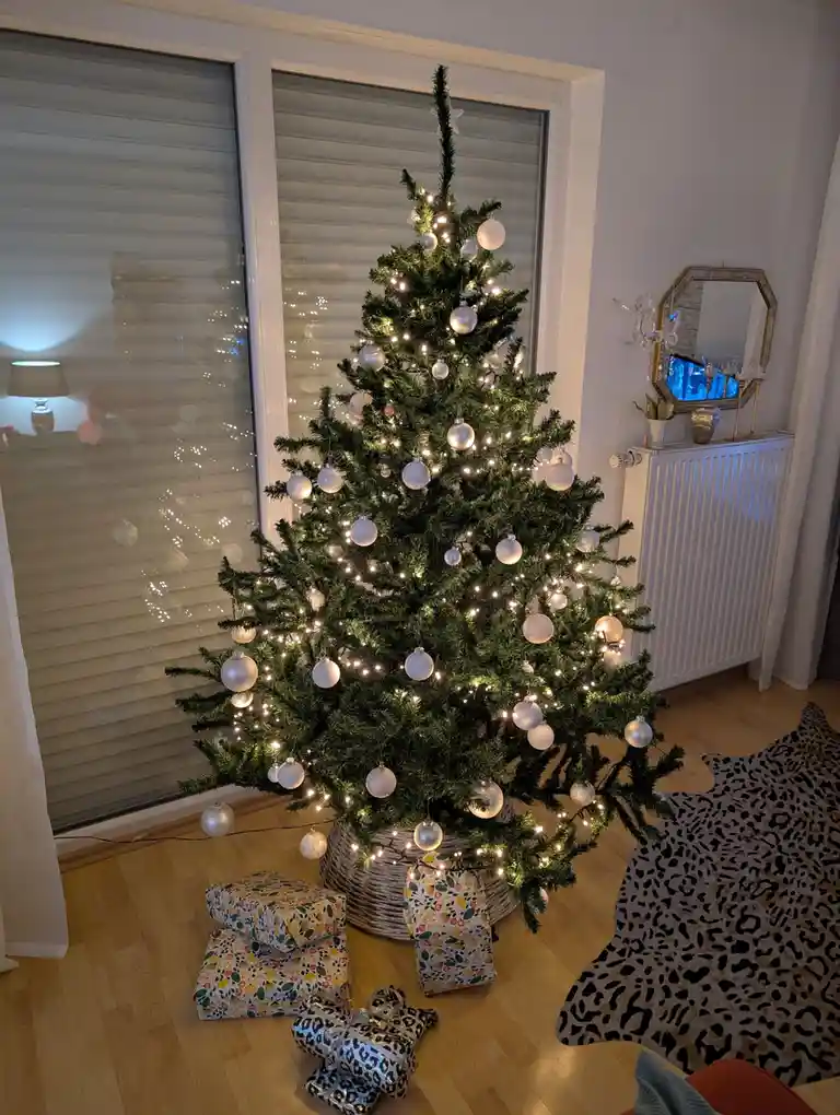 A decorated christmas tree with white ornaments and string lights stands in a room with wooden floors, next to a window with…