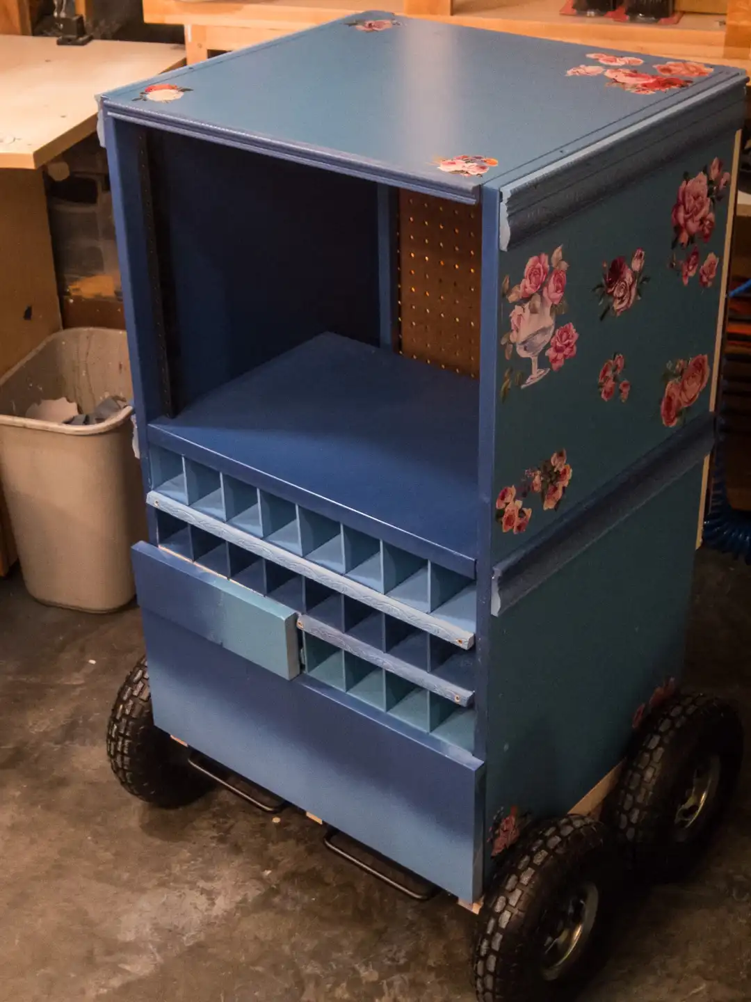 A blue, wheeled tool cart with floral decals and a pegboard interior is shown in a workshop. The cart has a top shelf, a…