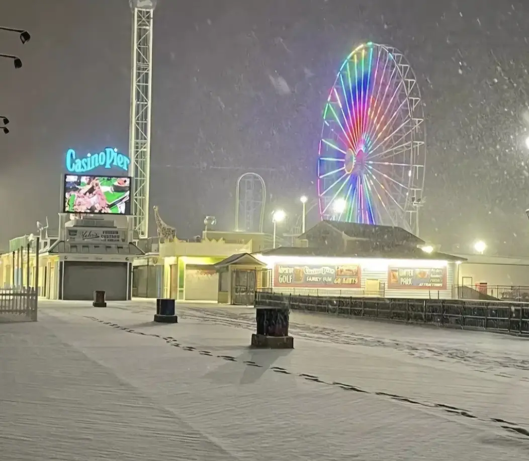a snowy night at the casino pier with a brightly lit ferris wheel and casino signage, featuring a trail of footprints…
