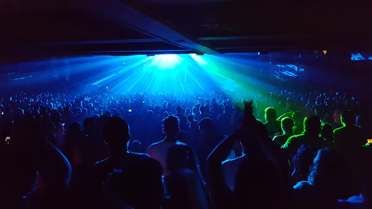 A crowded concert venue with a sea of silhouetted attendees facing a brightly lit stage bathed in blue and green laser…
