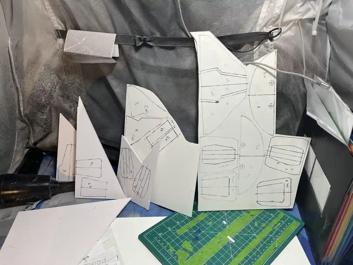 A cluttered crafting workspace shows multiple white paper patterns with black lines and handwritten markings, laid out on a…