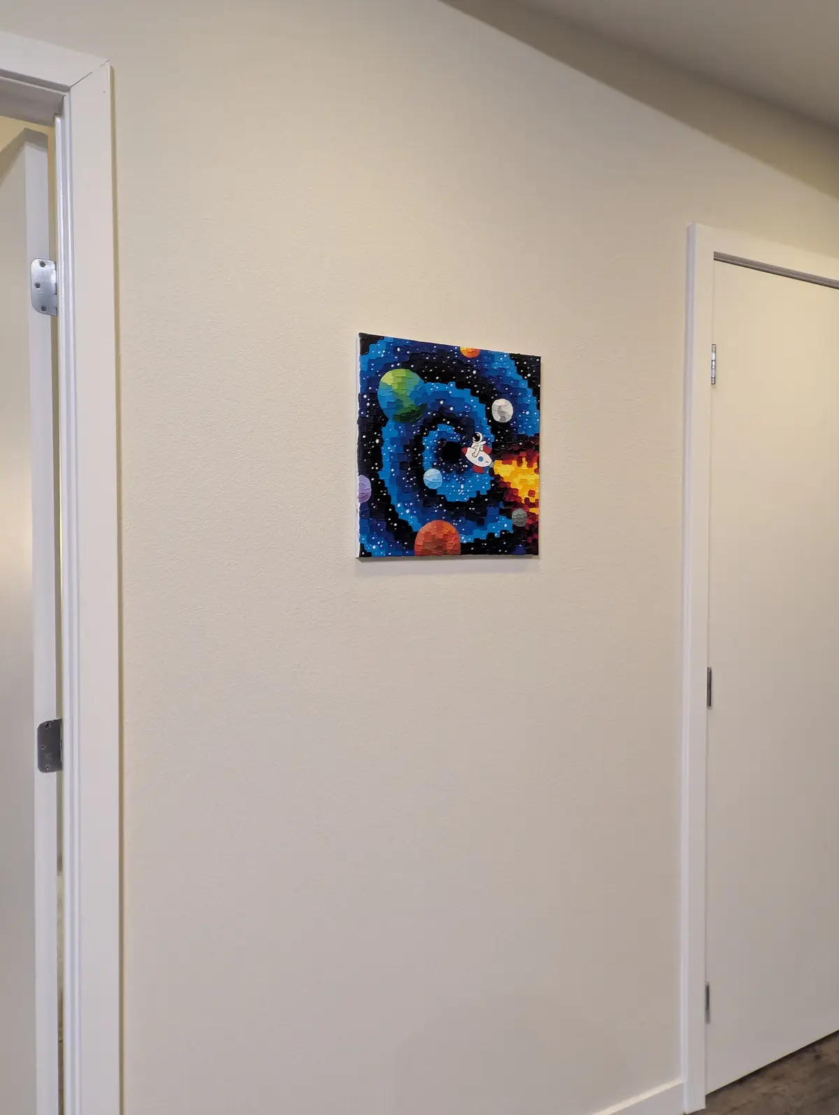 A small, colorful mosaic-style painting of a space scene with planets and a spaceship hangs on a plain beige wall between…