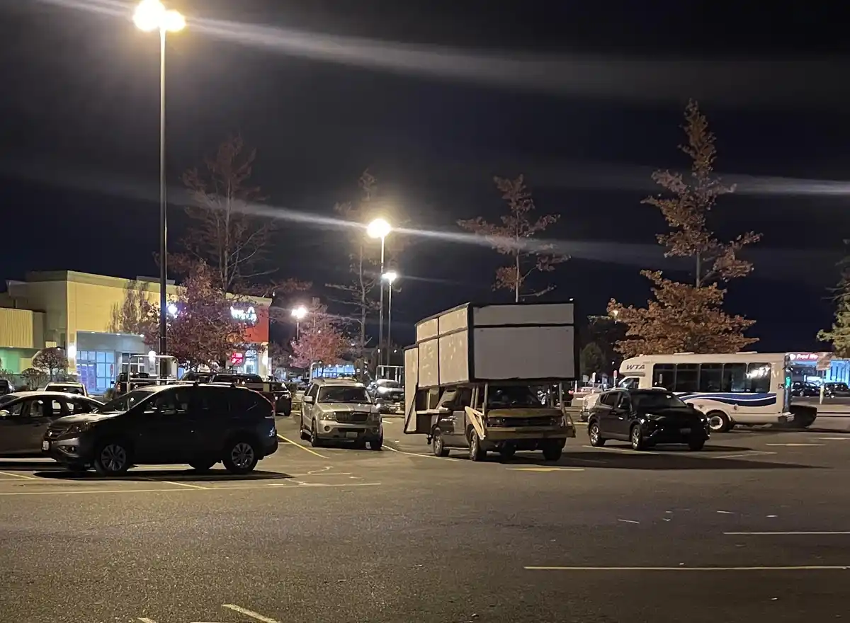 A nighttime photo of a parking lot with several cars and a bus, featuring a pickup truck with a large, boxy trailer mounted…