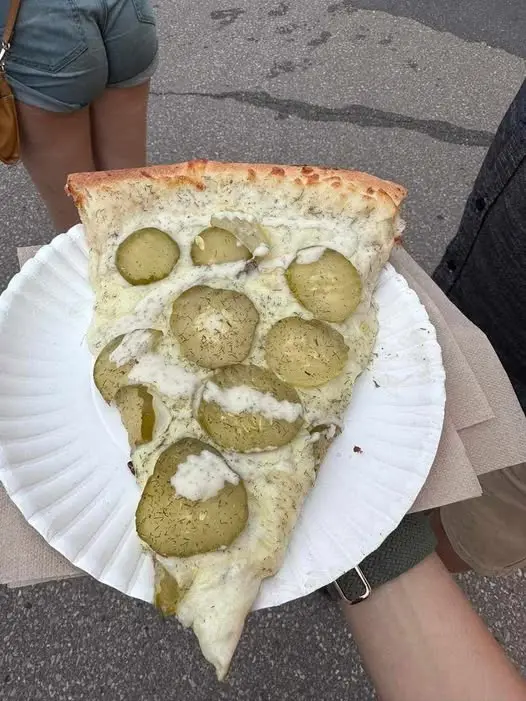A person holds a slice of pizza on a paper plate outdoors, topped with melted cheese and numerous round green pickles. In…