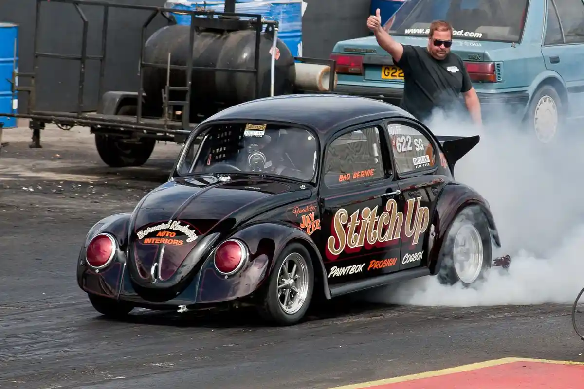 A black modified Volkswagen Beetle with 'Stitch Up' and 'Bad Bernie' decals is performing a burnout, kicking up thick white…