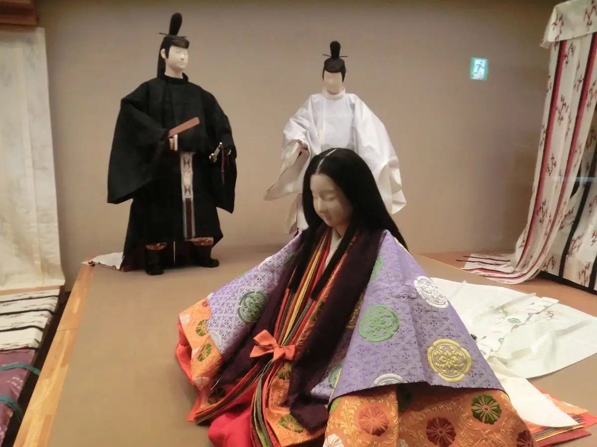 Three traditional Japanese mannequins dressed in formal ceremonial attire are displayed in a museum-like setting. The…