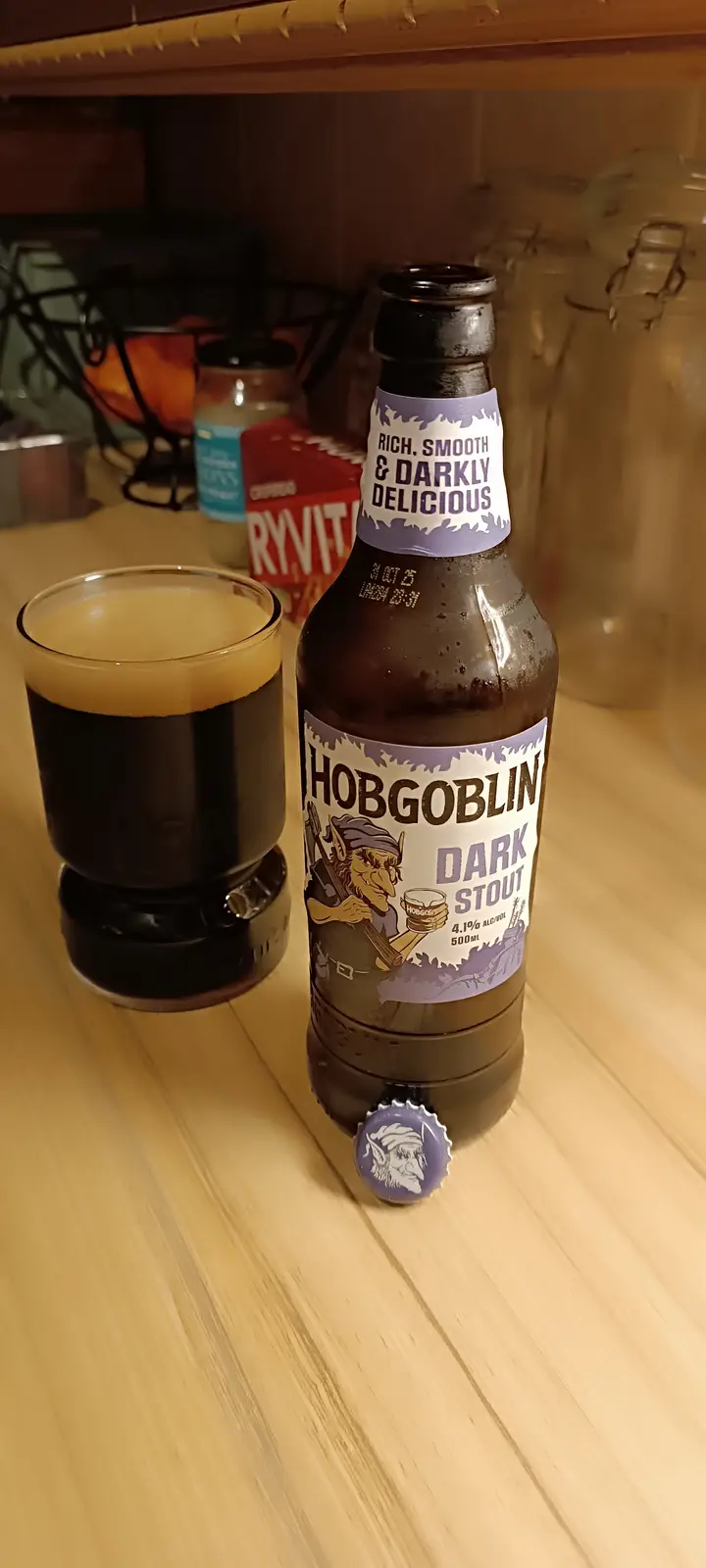 A bottle of Hobgoblin Dark Stout beer sits next to a glass filled with the dark beer, both resting on a wooden surface. The…