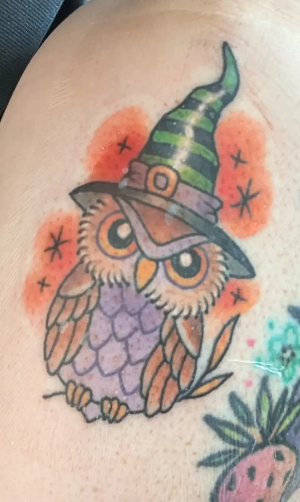 A colorful tattoo on skin shows a cartoon-style owl with large eyes, wearing a green and black striped witch's hat with a…