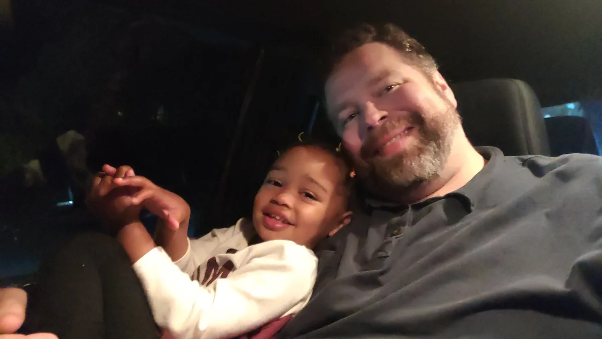 A man with a beard and a child are sitting in a car at night, smiling at the camera. The child is wearing a white shirt and…