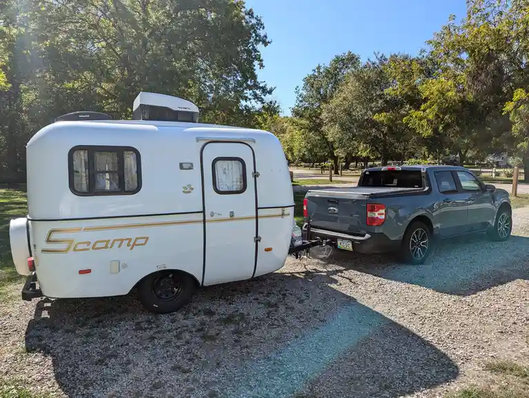 A white Scamp travel trailer with gold lettering is hitched to a gray Ford Maverick pickup truck, parked on gravel in a…