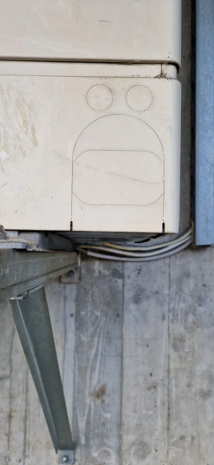 A close-up of a beige outdoor air conditioning unit mounted on a weathered wooden wall, showing faint pencil-like outlines…