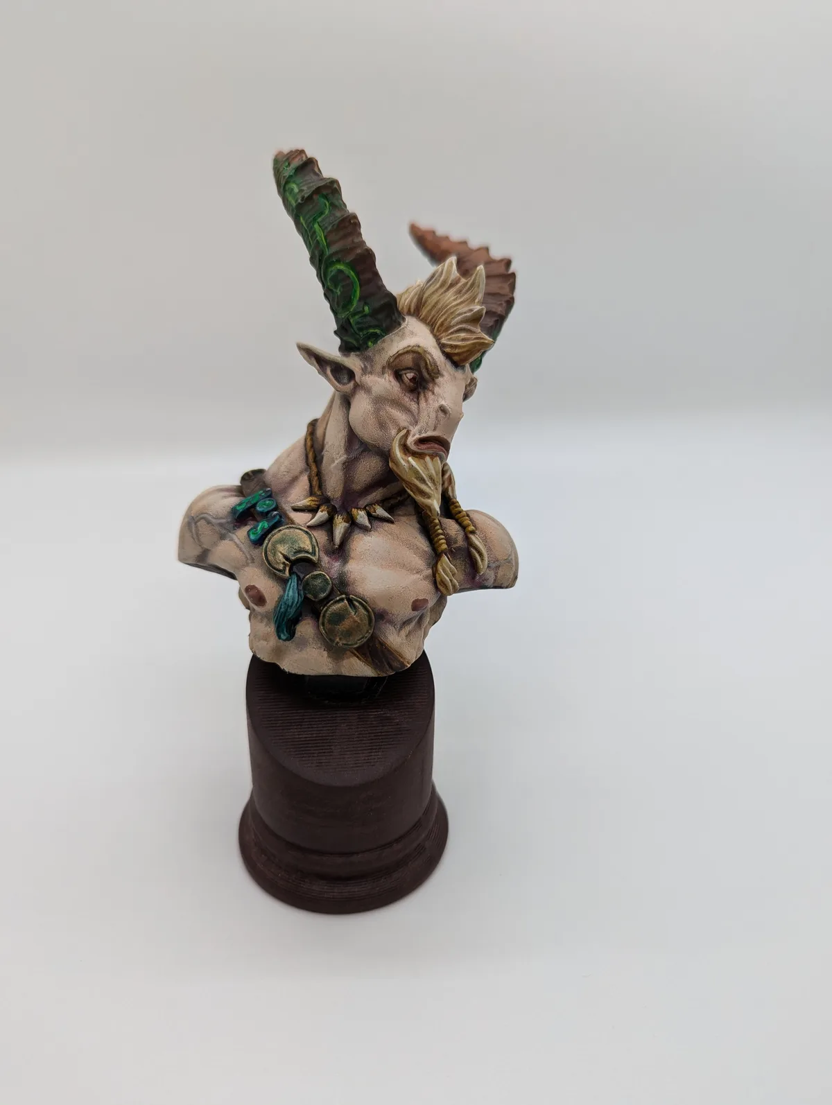 A painted fantasy bust of a horned humanoid figure with one large green-veined horn and one smaller brown horn, wearing…
