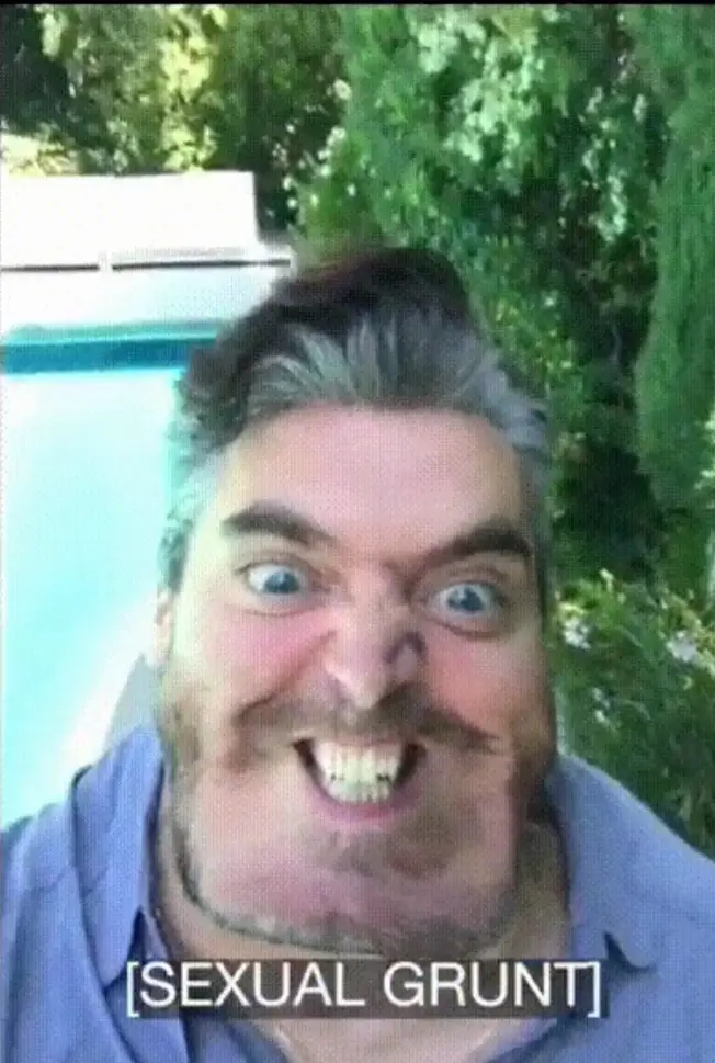 A man with a beard and gray hair makes a wide-eyed, toothy grin while wearing a blue shirt, standing near a pool with…