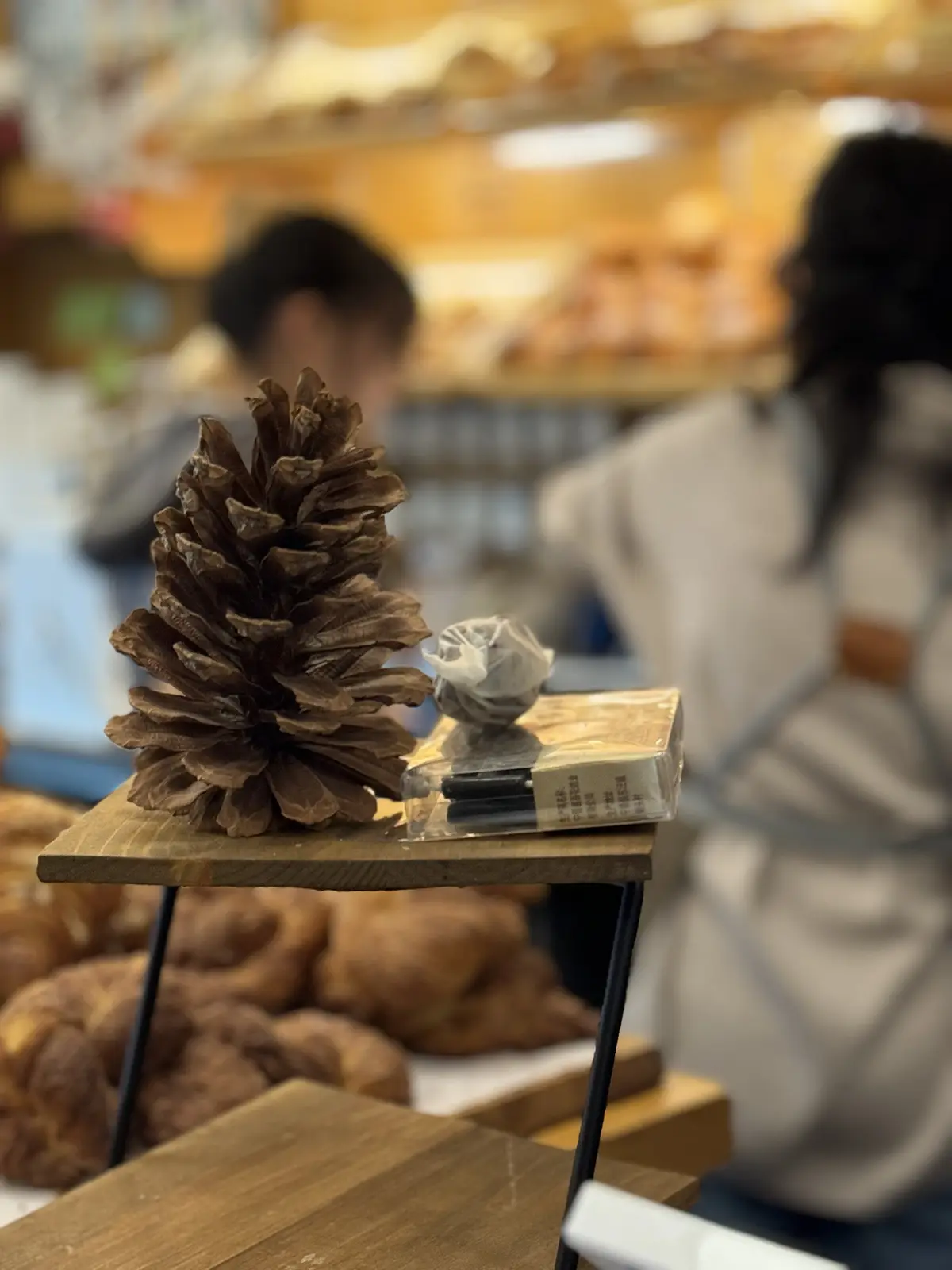 A large brown pinecone sits on a small wooden table next to a packaged item, with blurred people and pastries in the…