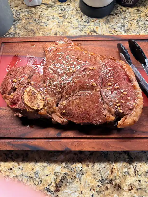 A large, cooked steak with a bone and seared crust sits on a wooden cutting board, glistening with juices and sprinkled with…