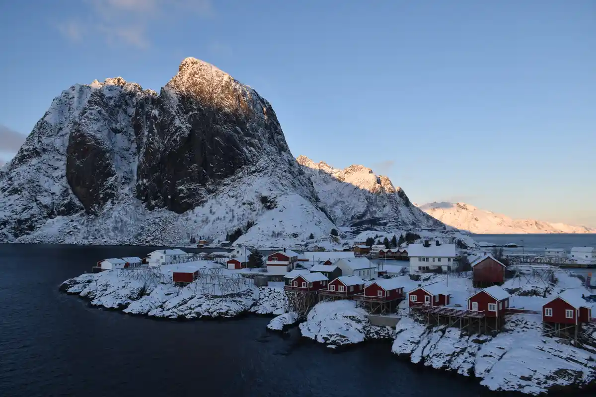 A scenic winter landscape shows a small village with red and white houses built on snow-covered rocky terrain beside a body…