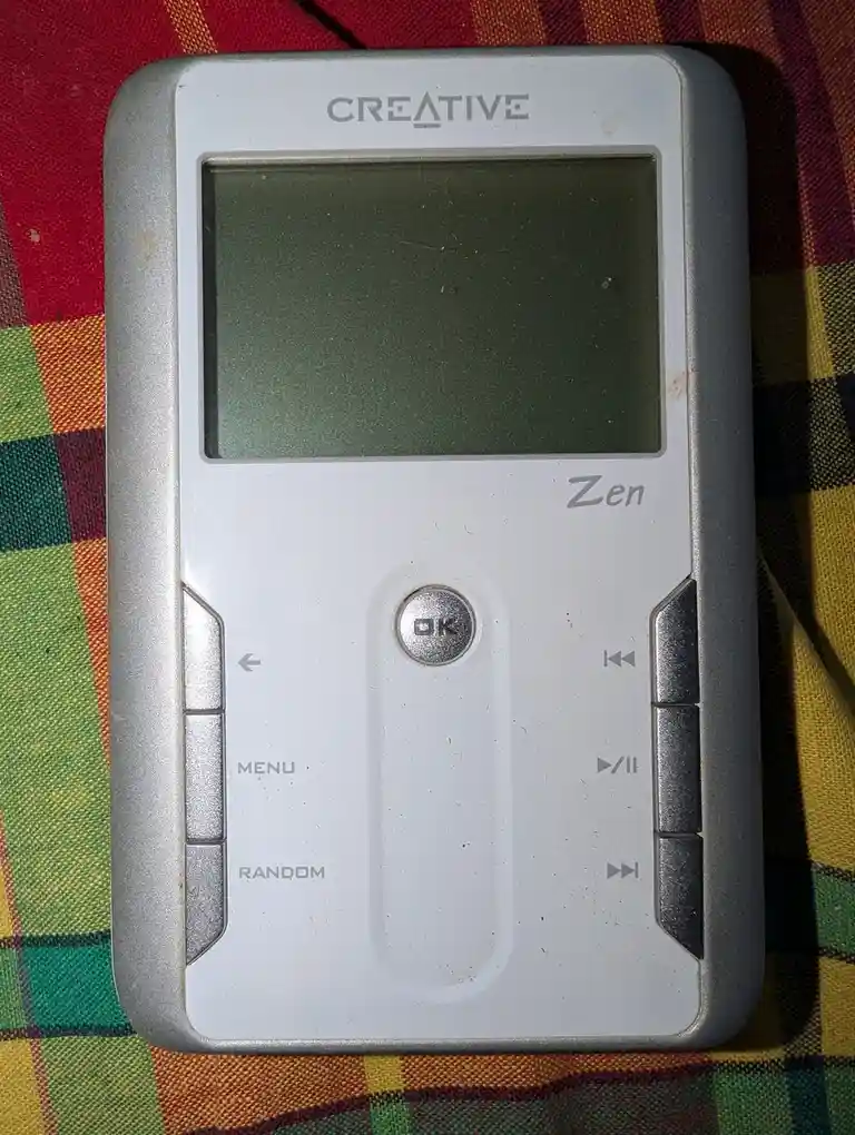 A close-up photo of a silver Creative Zen portable media player with a blank screen, resting on a colorful plaid fabric. The…
