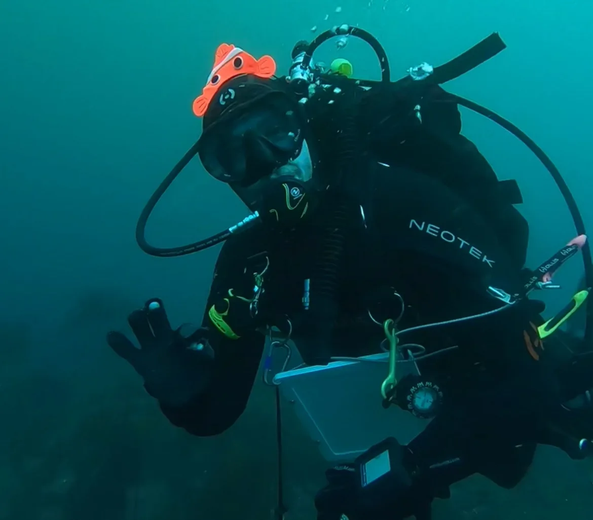 A scuba diver underwater wearing full gear including a mask, tank, and regulator, with a small orange clownfish plush toy…