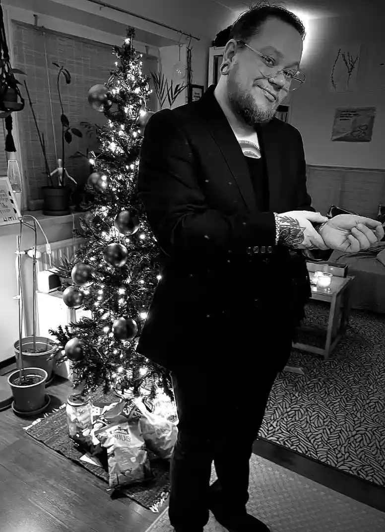 A black-and-white photo shows a man in a suit standing beside a lit Christmas tree, smiling while holding his hands open as…