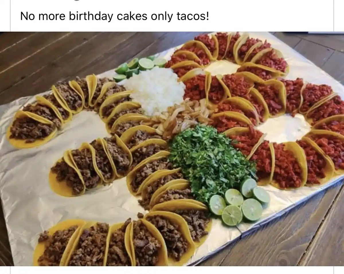 A large, elaborate arrangement of tacos and taco toppings is laid out on a foil-lined tray, forming the shape of a birthday…