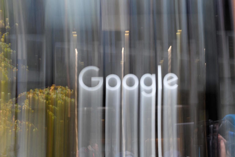 A blurred photograph of the Google logo on a glass storefront, with streaks of motion blur from passing pedestrians and…