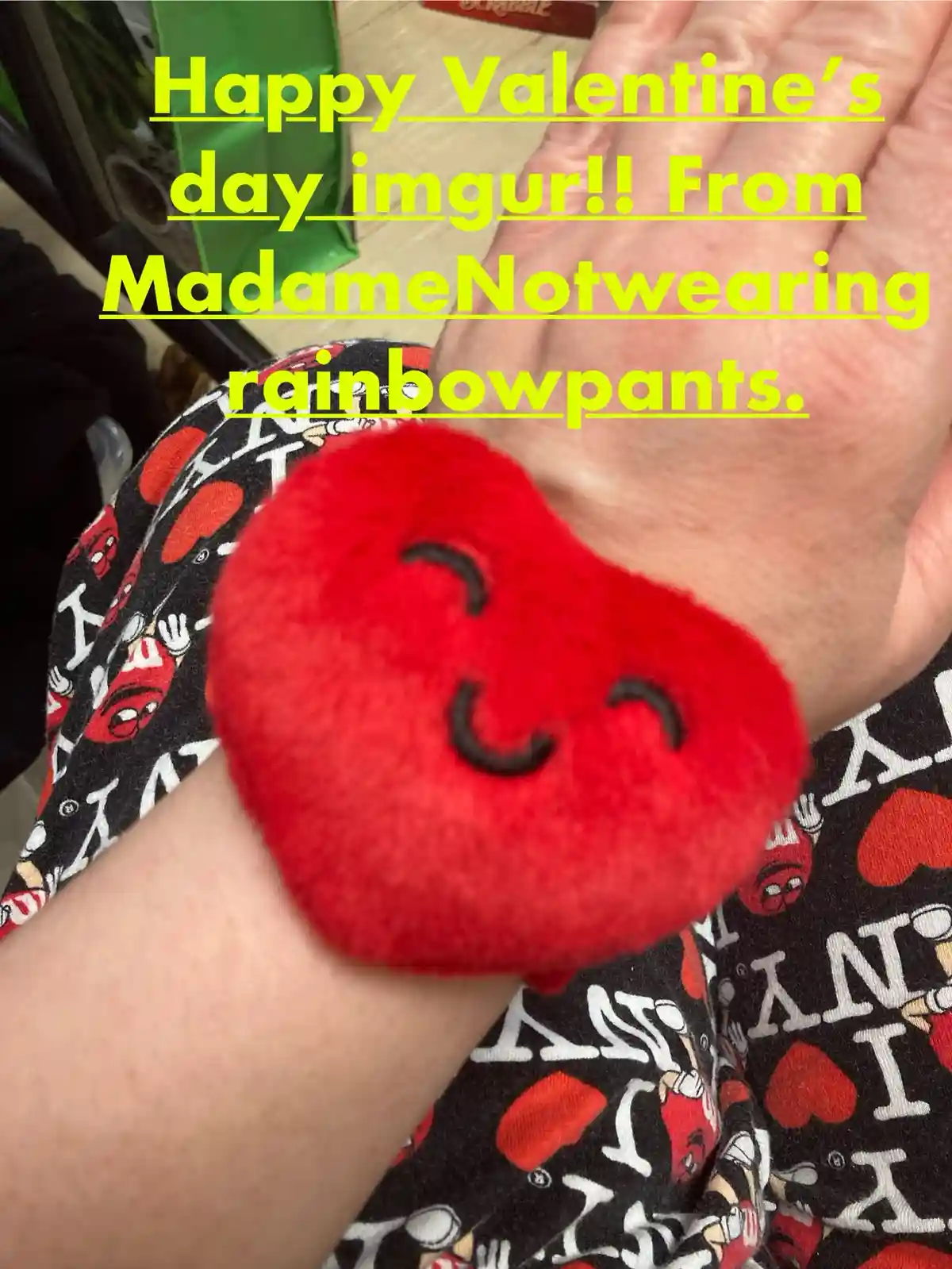 A close-up photo shows a person's wrist wearing a fluffy red heart-shaped wristband with a smiling face. The person is…