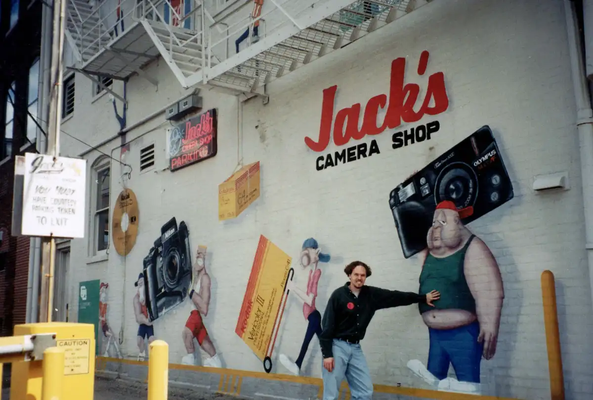 A man stands in front of a large mural on a building wall advertising 'Jack's Camera Shop'. The mural features cartoonish…