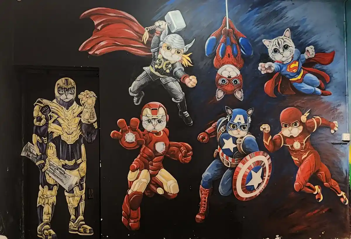 A colorful mural painted on a dark wall depicts seven anthropomorphic cats dressed as popular superheroes and villains…