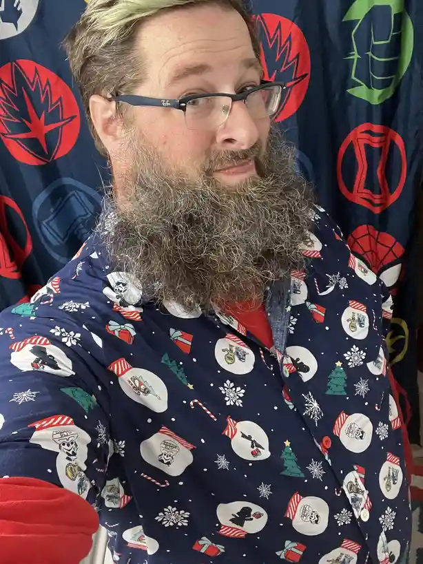 A man with a long gray beard and glasses wears a dark blue holiday-themed pajama shirt covered in snowmen, candy canes, and…