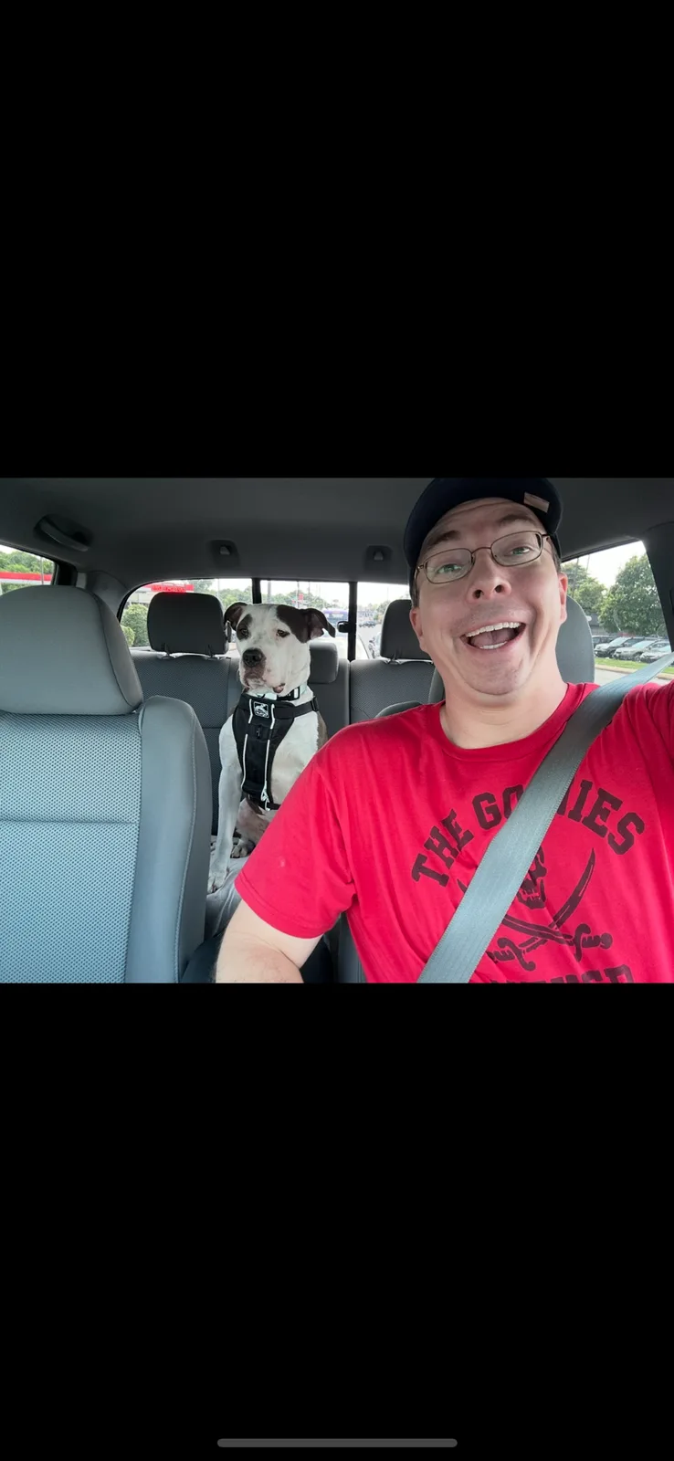 A man in a red t-shirt and glasses, smiling widely while driving, has a black and white dog wearing a harness sitting in the…
