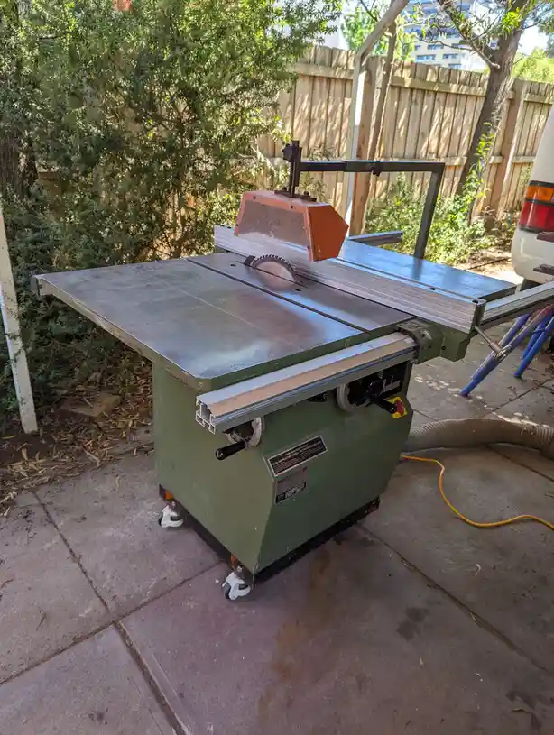 A green Woodman brand table saw sits outdoors on a concrete patio, surrounded by trees and a wooden fence, with a yellow…