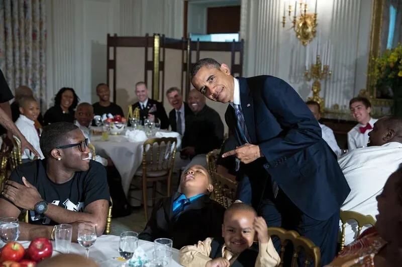 Barack Obama, in a dark suit, leans over a dining table and points at a young boy lying back with his eyes closed and a…
