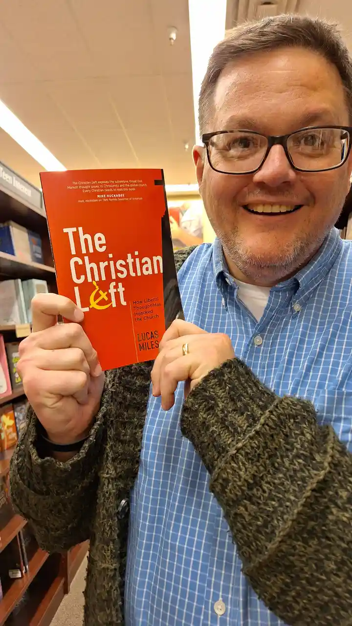 A smiling man in glasses and a blue plaid shirt holds up a red book titled 'The Christian Left' by Lucas Miles, where the…