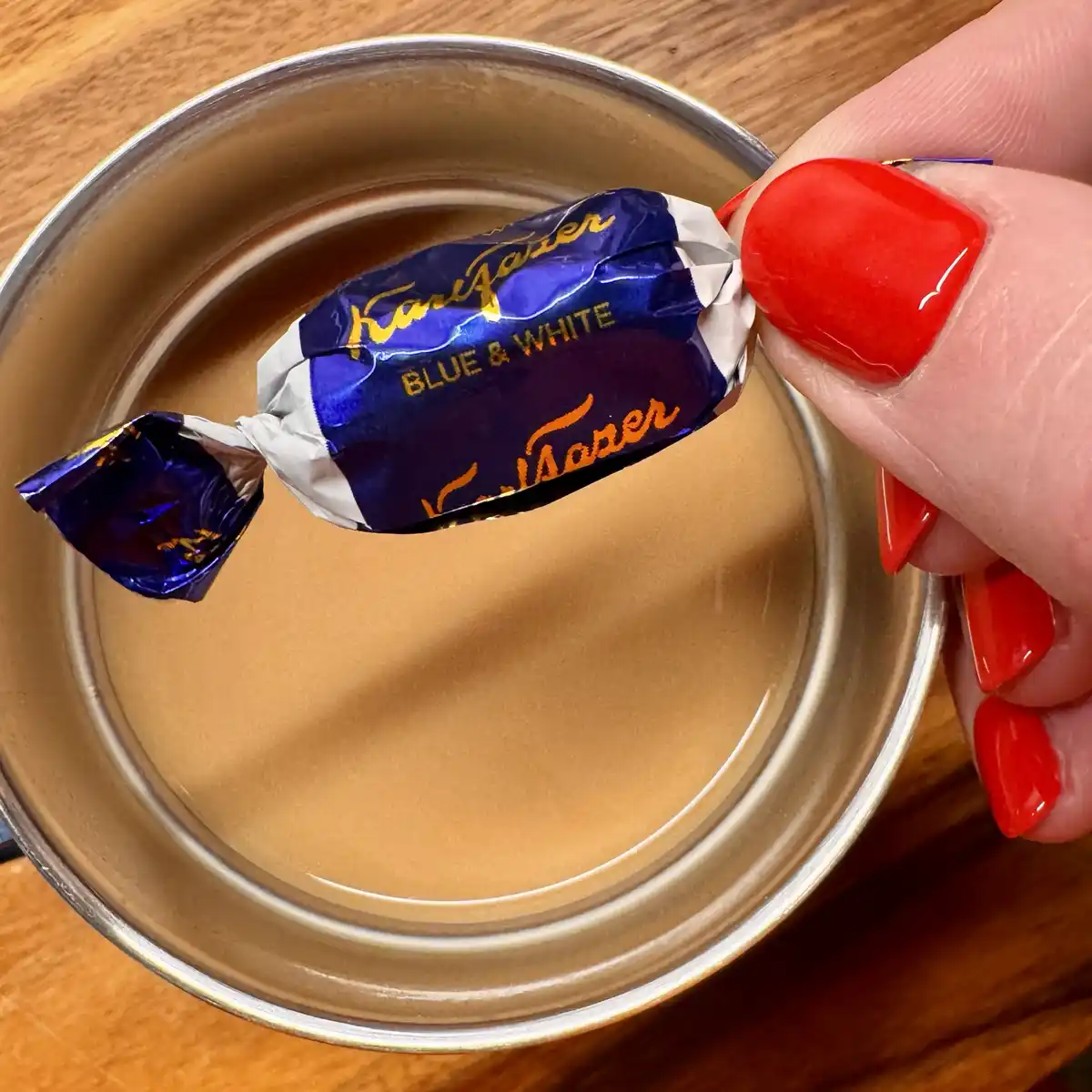 A hand with bright red nail polish holds a partially unwrapped Cadbury Blue & White chocolate candy over a glass of light…