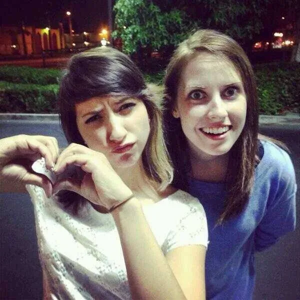 Two young women pose for a photo at night; the woman on the left makes a heart shape with her hands while scowling with a…