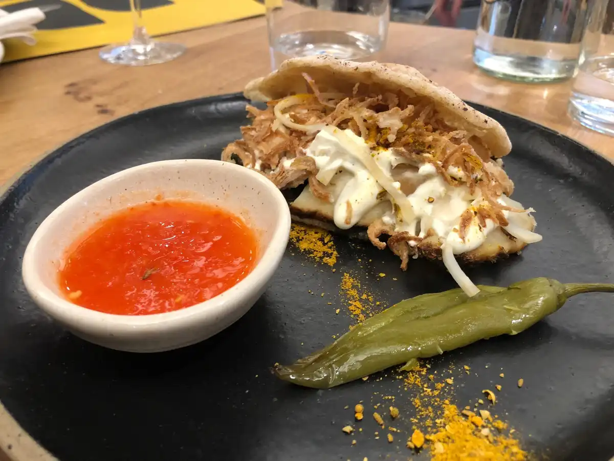 A close-up photo of a plated dish that looks like a sandwich with fried onions, a white sauce, and a pita-like bread, served…