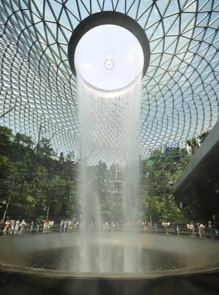 A wide-angle photo shows a massive indoor waterfall cascading from a circular opening in a glass-domed ceiling, surrounded…