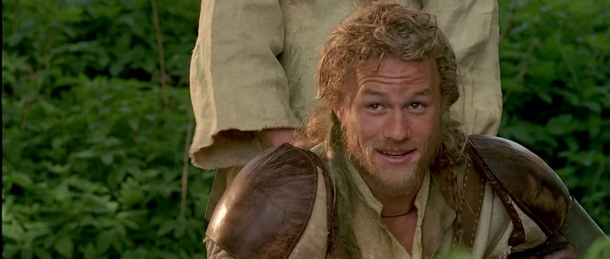 a man with curly hair and a beard, wearing medieval-style armor and a beige tunic, looks up with a slightly confused or…
