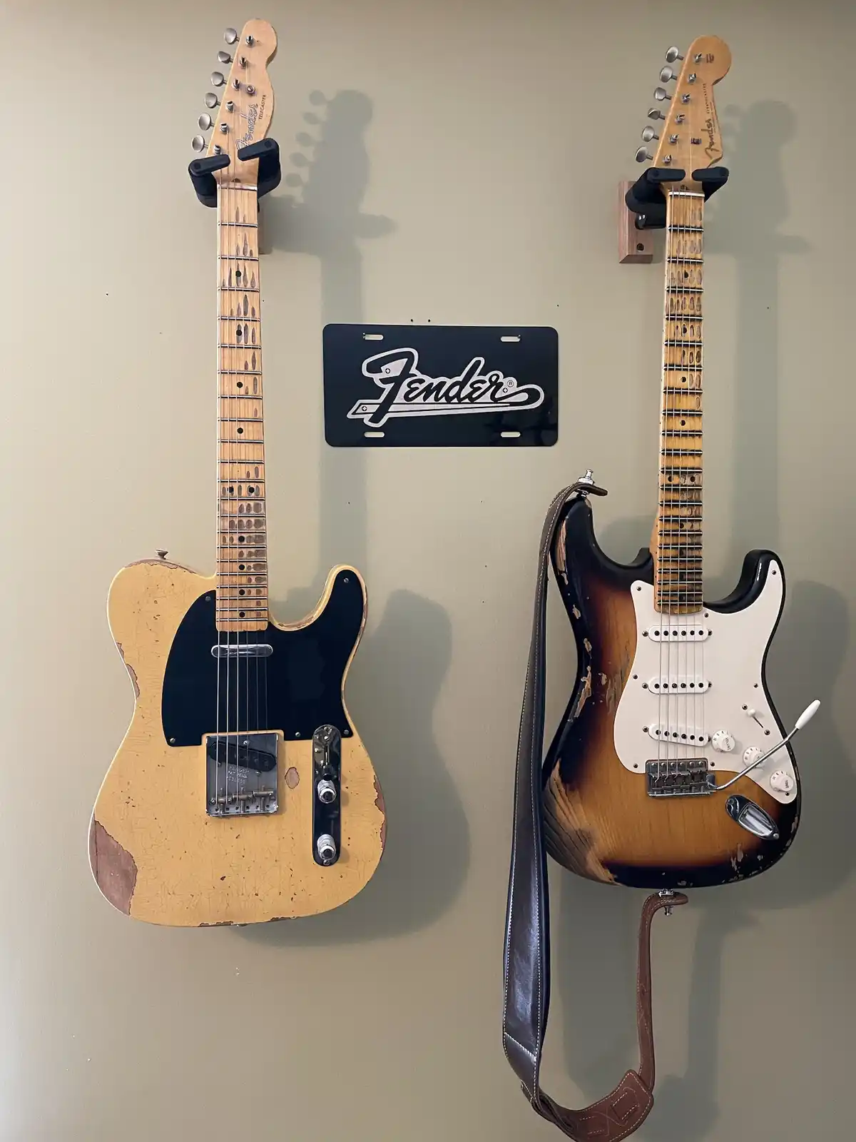 Two vintage, heavily worn Fender electric guitars — a Telecaster and a Stratocaster — hang vertically on a wall, flanking a…
