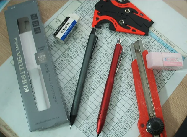 A collection of stationery items including a pencil sharpener, pens, a utility knife, and erasers arranged on a grid-lined…