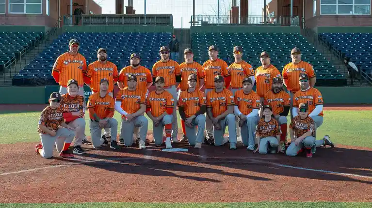 A baseball team poses for a group photo on a field, wearing orange jerseys with 'ST. LOUIS' printed across the chest. The…
